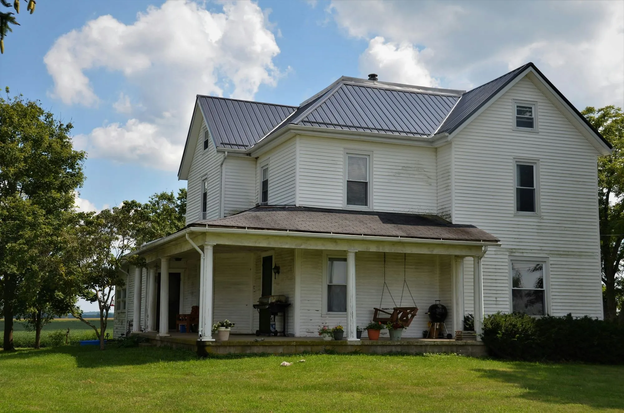 The finished exterior with new white siding, a rebuilt wraparound porch and matching brick chimney, set among mature trees and a green lawn